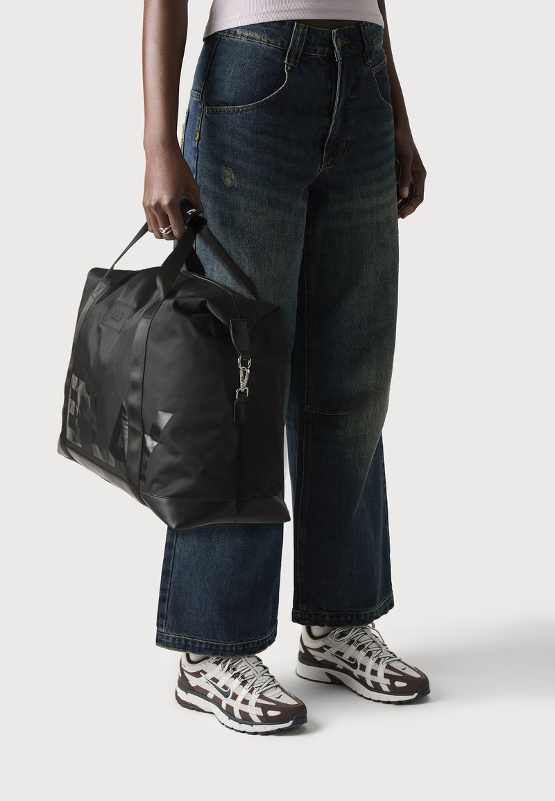 Borsa da viaggio nera realizzata in materiale sintetico con manici robusti, caratterizzata da una finitura lucida e dettagli con logo. Indossata con jeans a gamba larga in denim blu e scarpe.