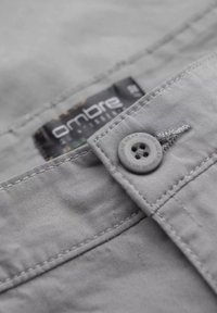 Tissu gris avec une texture lisse, comportant une fermeture à bouton et une étiquette indiquant "ombre". Des coutures sont visibles le long des bords.