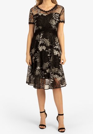 Kvinde iført en sort knælængde blondkjole med hvidt blomsterbroderi, gennemsigtige korte ærmer og sorte hæle med ankelrem, stående mod en hvid baggrund.