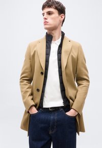 Νέος άνδρας με παλτό καμηλοπάρδαλη πάνω από μαύρο καπιτονέ μπουφάν και λευκό πόλο, με τα χέρια στις τσέπες των σκούρων μπλε τζιν.