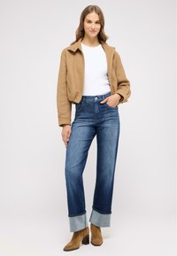 Braune Fleecejacke, weißes körperbetontes Top, hochgeschnittene blaue Jeans mit einem hellblauen Aufschlag. Braune Ankle Boots mit Blockabsatz.