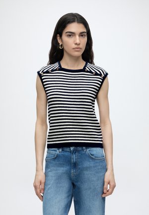 Ung kvinde med langt mørkt hår iført en navy og hvid stribet ærmeløs striktop med knapper, kombineret med blå denim jeans.