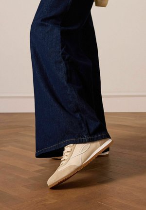 Beige athletic shoe with suede upper, lace-up design, and rubber sole. Worn with dark wide-leg jeans on a wooden floor.