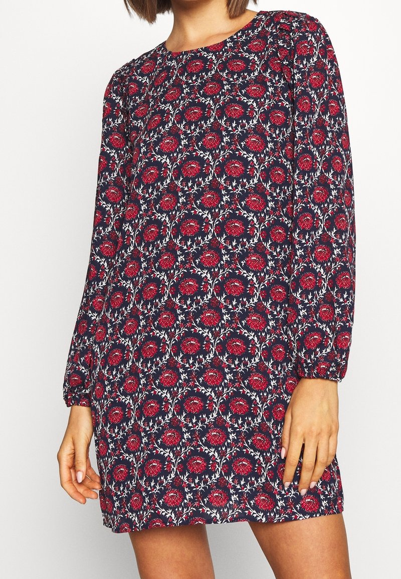 Femme portant une robe courte à manches longues avec un motif floral rouge sur fond bleu foncé, debout les mains détendues le long du corps.