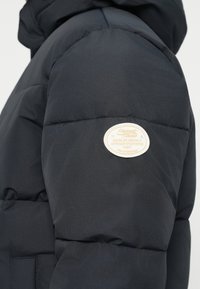 Mørkeblå pufferjakke med quiltede sektioner, der har et fremtrædende logo-patch i cremefarve påærmet. Glat, isoleret stof.
