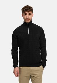 Musta neulepaita, jossa on korkea kaulus ja puolivälitasku vetoketjulla. Siinä on teksturoidun näköinen kuvio ja resorit hihansuissa. Päällä beiget housut.