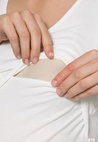 Kädet nostavat valkoista paitaa paljastaakseen vaalean beigeä liimalaastaria ihon päällä neutraalia taustaa vasten.