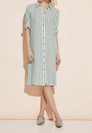 Vrouw draagt een knielange jurk met verticale groen-witte strepen, knoopjes aan de voorkant en opgerolde mouwen, gecombineerd met groene Mary Jane platte schoenen.