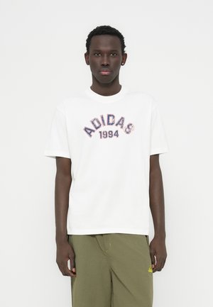 Fehér pamutpóló "ADIDAS 1994" logóval, piros és kék színben, kerek nyakú, rövid ujjú, olívazöld nadrággal viselve.