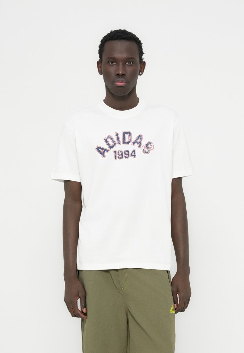 Λευκό βαμβακερό μπλουζάκι με το λογότυπο "ADIDAS 1994" σε κόκκινο και μπλε, με στρογγυλή λαιμόκοψη, κοντά μανίκια, φορεμένο με παντελόνι ελιάς.