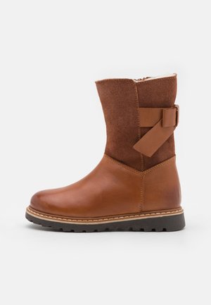 Botte marron mi-mollet avec partie inférieure en cuir lisse, dessus en suède, détail de bride latérale et semelle épaisse crantée sur fond blanc.