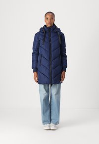 Doudoune marine avec motif en chevrons, présentant une fermeture éclair à l'avant, une capuche ajustable et des accessoires noirs. Associée à un pantalon flare en denim clair.