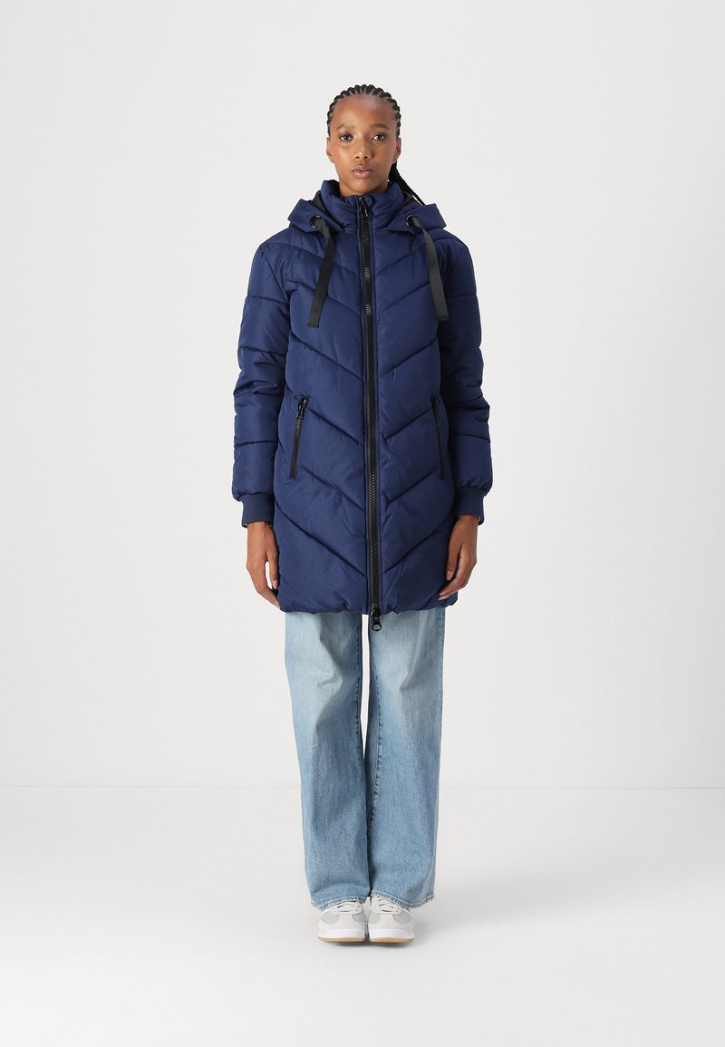 Doudoune marine avec motif en chevrons, présentant une fermeture éclair à l'avant, une capuche ajustable et des accessoires noirs. Associée à un pantalon flare en denim clair.