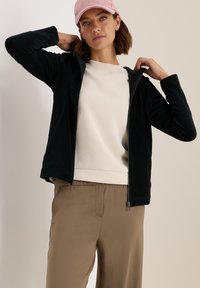 Haut à capuche zippé en velours côtelé noir avec poches latérales, assorti à un sweat-shirt beige clair et un pantalon kaki, porté avec une casquette rose clair.