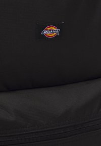 Mochila de tecido preto com uma textura suave, apresentando um patch do logo da Dickies em vermelho e amarelo. Inclui um detalhe de zíper visível.