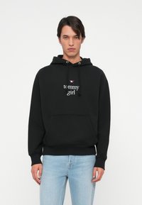 Mladý muž v čiernej mikine s kapucňou s textom a logom "tommy girl", v kombinácii s svetlomodrými džínsy, stojí pred čistým bielym pozadím.