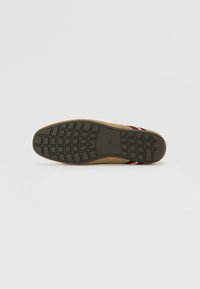 Suela de zapato de ante en color marrón claro con una parte inferior de goma negra texturizada que presenta un patrón de rejilla. Acentuado con una correa trenzada en rojo y negro.