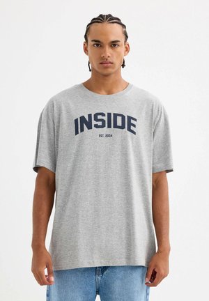 Camiseta de algodón gris con corte holgado, cuello redondo y "INSIDE EST. 2004" impreso en azul marino en el pecho. Estilo informal.