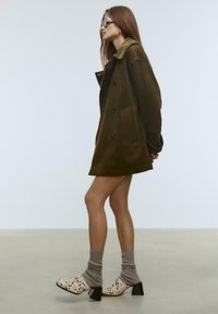 Khakifarbener Cordmantel mit großen Knöpfen, getragen über einem braunen Strickkleid. Gemusterte Absatzschuhe mit schwarzen Sprenkeln und grauen Socken.