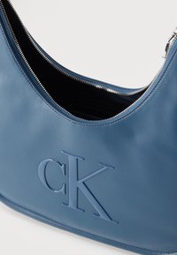 Μπλε δερμάτινη τσάντα χειρός με ασημένιο φερμουάρ και ανάγλυφο λογότυπο CK, μισάνοιχτη για να αποκαλύψει το μαύρο υφασμάτινο εσωτερικό.