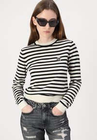 Stripet strikkegenser i svart og krem, med lange ermer og ribbestrikkede mansjetter, kombinert med slitt svarte jeans. Lite logo vist.