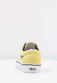 Baskets Vans en daim jaune avec semelle blanche et bande noire, vues de dos sur une surface et un fond blancs.