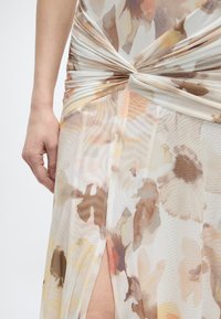 Κοντινό πλάνο ενός χεριού που ακουμπά δίπλα σε ένα διαφανές φόρεμα με λουλουδένιο σχέδιο, με έναν στριφογυριστό κόμπο και μια εμπρόσθια σχισμή που αφήνει να φαίνεται δέρμα από κάτω.