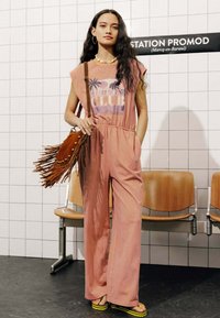 Combinaison rose à manches courtes, taille élastique et motif de palmiers. Accessoirisée d'un sac à franges marron et de sandales plateformes avec des accents jaunes.