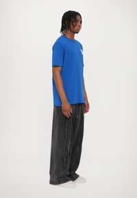 Blue cotton T-shirt with short sleeves and a logo on the left chest, paired with loose-fitting black denim pants and light shoes.