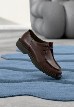 Mocassin en cuir marron avec semelle noire flottant au-dessus d'un tapis bleu texturé, avec une sculpture abstraite grise en arrière-plan.