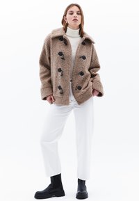 Beige fleece takki mustilla napilla, iso kaulus ja löysä istuvuus. Päällä valkoinen poolopaita ja yhdistetty valkoisiin housuihin.