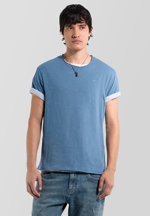 Giovane uomo che indossa una t-shirt blu con maniche arrotolate, jeans in denim e una collana nera con piastrina militare, in piedi davanti a uno sfondo chiaro e uniforme.
