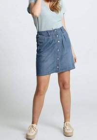 Gonna in denim con vita alta, chiusura con bottoni anteriori, pieghe e tasche laterali. Indossata con una maglietta a maniche corte texturizzata di colore azzurro chiaro e scarpe beige.