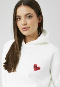 Sweat à capuche blanc à texture pelucheuse, avec un patch cœur rouge orné d'un texte brodé. La capuche est surdimensionnée, renforçant le design décontracté.