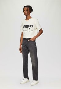 Biała koszulka z grafiką z napisem "SOUNDTRACK", w połączeniu z ciemnoszarymi dżinsami i białymi trampkami, stojąca na neutralnym tle.