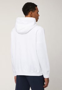 Haut en coton blanc avec une capuche spacieuse, des poignets et un ourlet côtelés. Texture lisse, coupe décontractée, détails de couture visibles au dos.