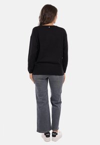 Donna in piedi di spalle, indossando un top nero a maniche lunghe, jeans grigi dritti e sneakers bianche con il tallone nero.