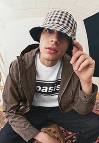 Brun zip-up jakke, hvid T-shirt med sort "oasis" logo, mørke denim jeans og ternet bucket hat i blå og brune nuancer.