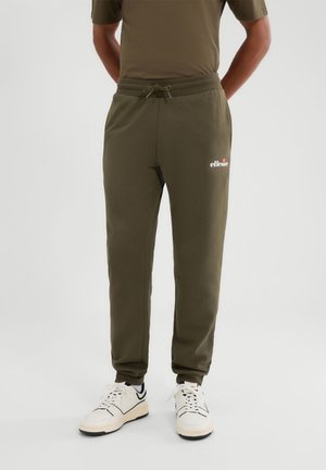Man wearing olive green ellese sweatpants and white sneakers standing against a plain light background.