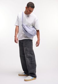 Borsa a tracolla di un leggero colore viola, realizzata in tessuto, indossata a tracolla. T-shirt bianca casual, jeans larghi grigio scuro e sneakers bianche con accenti blu.