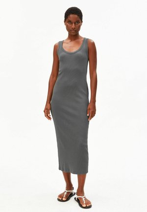 Frau steht, trägt ein ärmelloses, figurbetontes graues Midikleid und schwarze flache Sandalen mit weißen Riemen vor einem schlichten weißen Hintergrund.