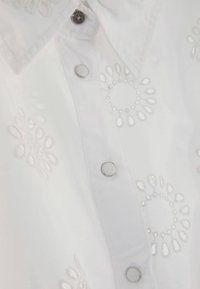 Chemise blanche avec boutons-pression métalliques ronds et motifs de broderie florale découpée près du col et de la patte de devant.