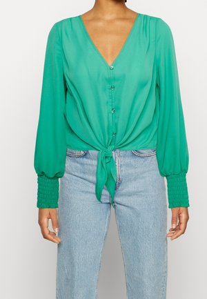 Femme portant un chemisier vert vif à boutons avec de longues manches bouffantes et un nœud à l'avant, associé à un jean bleu clair taille haute.
