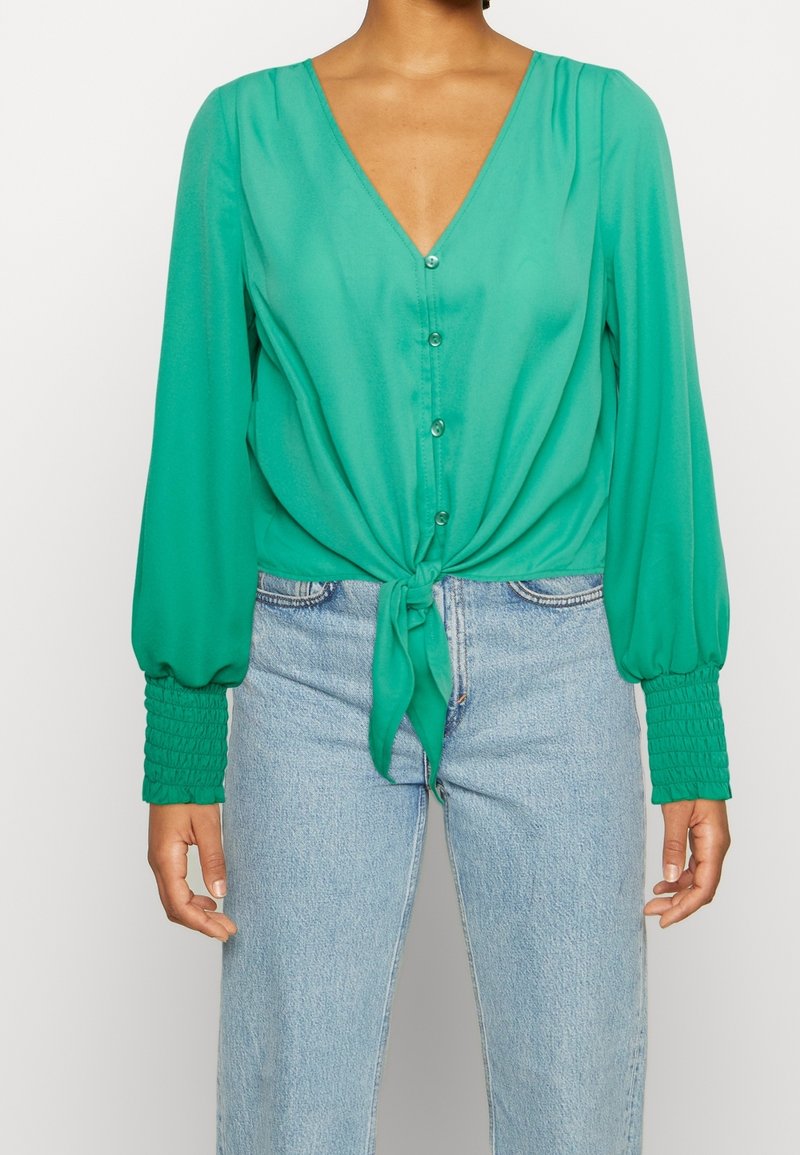 Femme portant un chemisier vert vif à boutons avec de longues manches bouffantes et un nœud à l'avant, associé à un jean bleu clair taille haute.