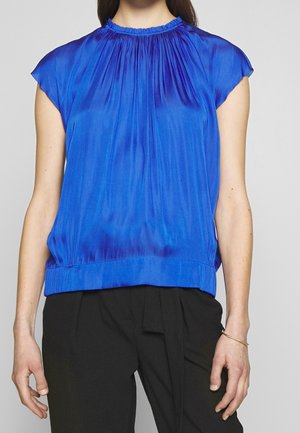 Vrouw die een mouwloze, koningsblauwe geplooide blouse draagt, gecombineerd met een zwarte broek met hoge taille, staand tegen een grijze achtergrond.