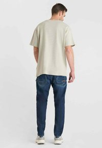 Man die met zijn rug naar de camera staat, gekleed in een beige T-shirt met korte mouwen, donkerblauwe jeans en lichte sneakers, tegen een effen achtergrond.