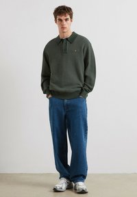 Zielony sweter prążkowany z kołnierzykiem, zapięciem na guziki i małym logo, w zestawieniu z luźnymi niebieskimi dżinsami i srebrnymi trampkami.