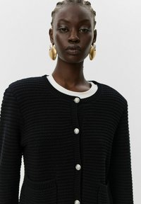 Črn rebrast cardigan z belim ovratnikom, ki ima srebrne gumbke in stranske žepke. Gladka tekstura, zasnova z gumbi in moderna krojenje.