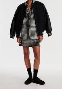 Musta yliopistojakku, jossa on nahkahihat, houndstooth-kuvioinen bleiserimekko, mustat sukat ja mustat sujautettavat kengät, joissa on hihnat.