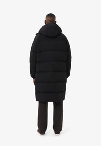 Abrigo negro acolchado con capucha, de corte largo y diseño acolchado horizontal. Se lleva sobre pantalones oscuros, visto desde atrás.